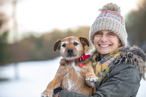 A portrait of a mature woman wearing warm clothing, holding her senior patterdale terrier while standing outdoors in the snowy Northumberland weather. They are both looking at the camera and the woman is smiling.