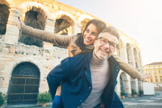 casal de idosos felizes apaixonados por se divertir em frente ao famoso monumento da itália. visão de baixo ângulo de mulher alegre esticando os braços enquanto senta na parte de trás de seu marido - férias, turismo e conceito de viagem - italy coliseum rome italian culture - fotografias e filmes do acervo