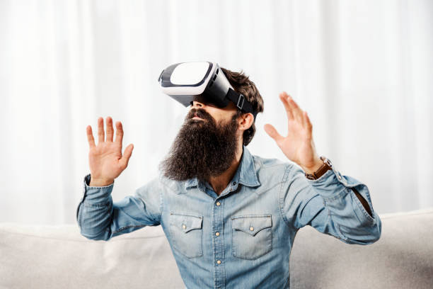 A young bearded man having VR headset and having virtual reality experience at home.