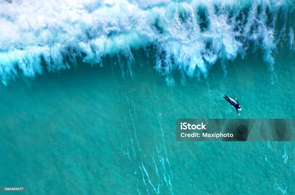 Big wave splashing behind a lonely surfer seen from above Big sea wave splashing with foam while a lonely surfer wearing a diving suit is seen from above swimming to calm waters. Ocean view from above, drone point of view. Surfing Stock Photo