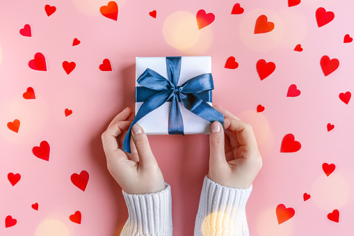 Female Hands in sweater holding a gift in white wrapping paper on pink background with red hearts. St. Valentines Day, love, tenderness, friendship and care concept. Cozy, festive, romantic wallpaper