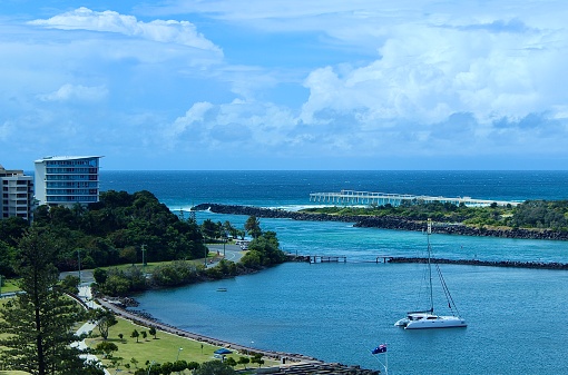 Horizontal landscape high up aerial view looking down to Tweed Heads River with view out to blue aqua turquoise ocean with promenade jetty yacht boat public park with Australian flag and high rise apartment buildings from window