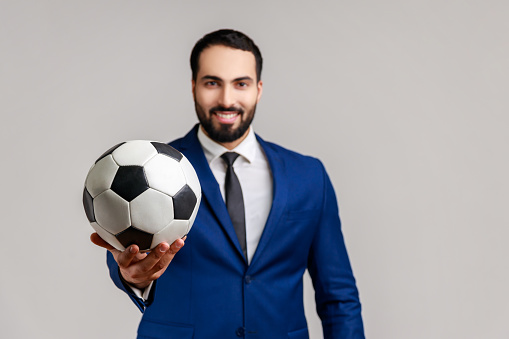 Happy attractive bearded businessman holding out soccer ball on his hand with smiling optimistic expression, wearing official style suit. Indoor studio shot isolated on gray background.