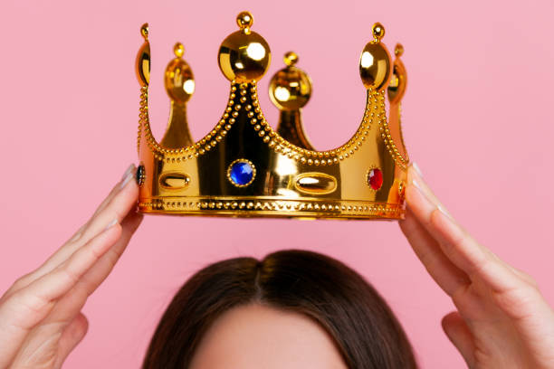 Unknown brunette woman putting on golden crown, arrogance and privileged status. Unknown woman putting on golden crown, arrogance and privileged status, concept of self confidence in success, self-motivation and dreams to be best. Indoor studio shot isolated on pink background. showing off stock pictures, royalty-free photos & images