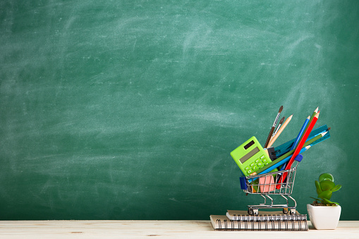 Education concept - school supplies in a shopping cart on the desk in the auditorium