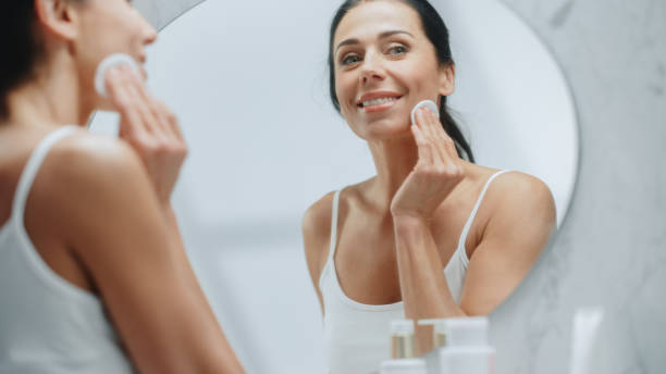 Beautiful Caucasian Woman Uses Cotton Pad to Clean Face Makeup, Skin, Smiles in the Mirror. Happy Middle Aged Female Enjoying Her Beauty Routine. Wellness, Natural Beauty, Cosmetic Skincare Products hermosa mujer caucásica usa algodón para limpiar el maquillaje de la cara, la piel, las sonrisas en el espejo. feliz mujer de mediana edad disfrutando de su rutina de belleza. bienestar, belleza natural, productos cosméticos para el cuidado de la piel