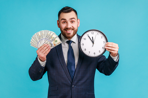 Portrait of happy businessman wearing official style suit showing dollar cash and wall clock on camera, hourly payment, looking at camera. Indoor studio shot isolated on blue background.