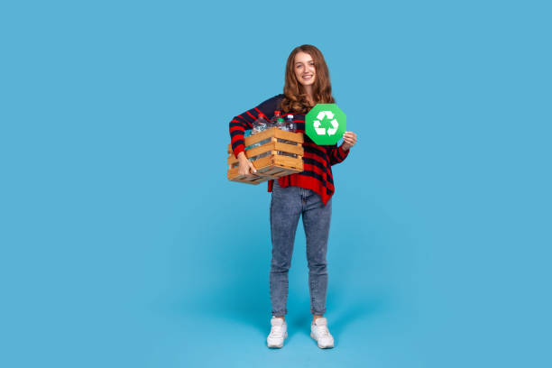 Woman holding box with empty plastic bottles and green recycling sign, fights environmental issues. Full length portrait of happy woman in striped sweater, holding box with empty plastic bottles and green recycling sign, fights environmental issues. Indoor studio shot isolated on blue background. recycle symbol cut out stock pictures, royalty-free photos & images