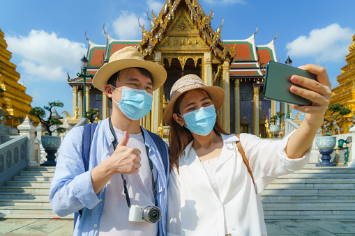 Asian couple happy tourists to travel wearing mask to protect from Covid-19 on they holidays and selfie by camera in Wat Phra Kaew Temple in Bangkok, Thailand