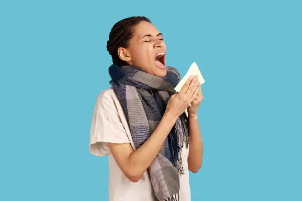 Sick woman with black dreadlocks feels unwell, sneezing in tissue, suffers from running nose, cold symptoms or allergy, wearing white shirt. Indoor studio shot isolated on blue background.