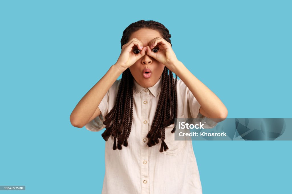 Woman covering eyes with ok signs, makes binoculars, foolishes around and looks through goggles. Funny playful woman with black dreadlocks covering eyes with ok signs, makes binoculars, foolishes around and looks through goggles, wearing white shirt. Indoor studio shot isolated on blue background Peeking Stock Photo