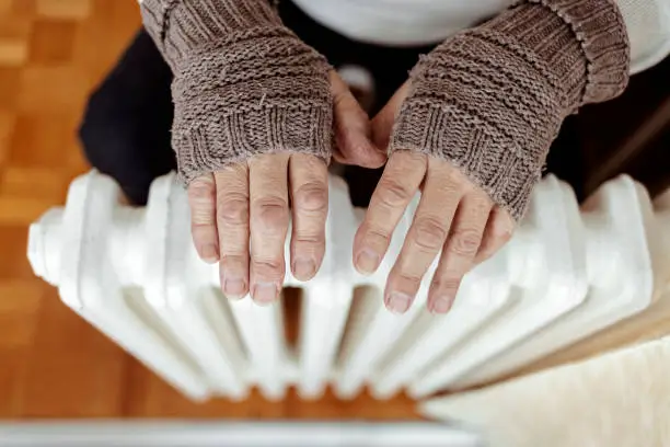 A man hands in wool gloves warm near the heater. Old men's hands in knitted gloves on heating radiator at home during the day. Person heating their hands at home over a domestic radiator in winter.