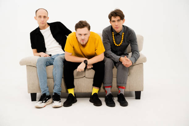 istock Young Guys Sit on a Sofa on a White Background and Look Together at the Camera. Three Friends are Sitting on the Couch and Watching TV, Watching a Home Theater Together. Close-up 1364607923