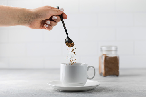A woman adds instant coffee to a white mug on grey stone table in light kitchen closeup