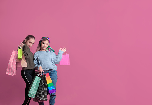 Positive friendly 11 year old girls standing with many paper shopping bags against pink background and looking at camera in studio