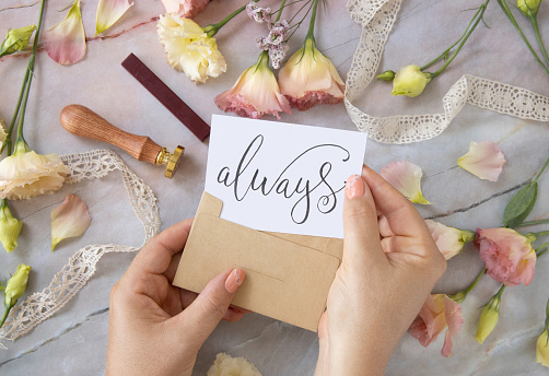 Hands with Card ALWAYS inside envelope near by pink flowers, wax seal and ribbons on a marble table top view. Romantic scene with handwritten card flat lay. Valentines concept