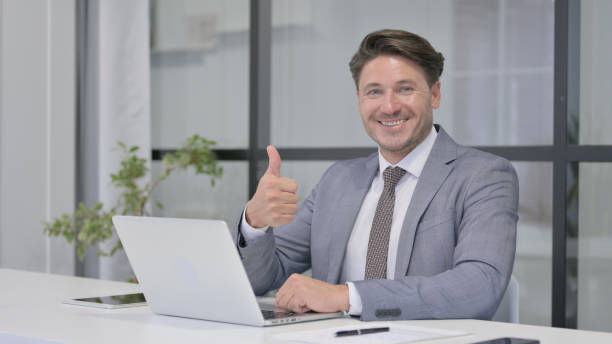 istock Middle Aged Man Showing Thumbs Up Sign While using Laptop at Work 1364617695