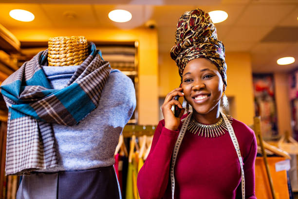 mujer tanzana con turbante con estampado de serpiente sobre escuchar trabajar en tienda de telas llamando al cliente por teléfono inteligente - tienda de telas fotografías e imágenes de stock