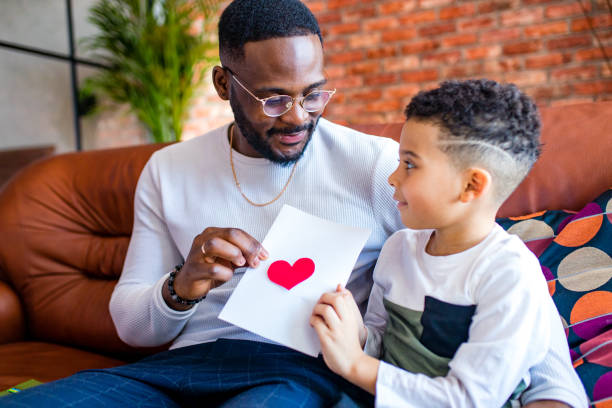 afro americano bebê dando ao pai uma foto de dia dos namorados na sala de estar - fathers day greeting card father child - fotografias e filmes do acervo