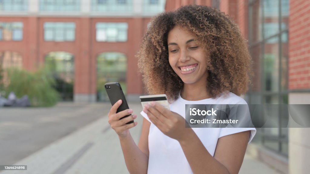African Woman Shopping Online on Smartphone, Outdoor Portrait of Young African Woman making Online Payment on Smartphone Credit Card Stock Photo