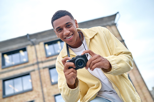 Right moment. Bottom view of friendly dark-skinned man with toothy smile holding photo camera in hands looking at camera against background of building and sky on fine day