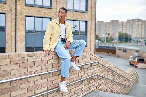 Nice location. Young dark-skinned man in casual light clothes with camera around his neck sitting on brick wall on city street contented looking to side