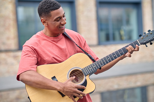 Musician. Dark-skinned young adult smiling man in red t-shirt playing guitar standing against city building background on fine day