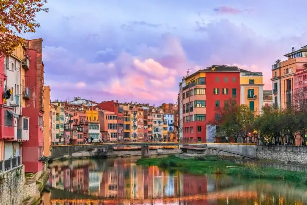 Evening landscape of the Old Town of Girona with pink clouds over Pont den Gomez o de la princesa bridge, Spain. Bright colorful buildings are reflected in the water of the Onyar River