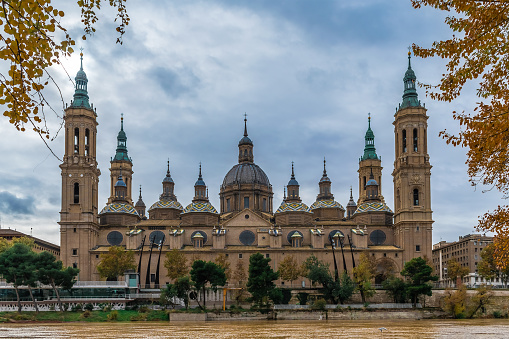 Cathedral-Basilica of Our Lady of the Pillar on the embankment of Ebro river with yellow water in Zaragoza on an autumn day, Spain. Panoramic view of the Spanish Roman Catholic Church