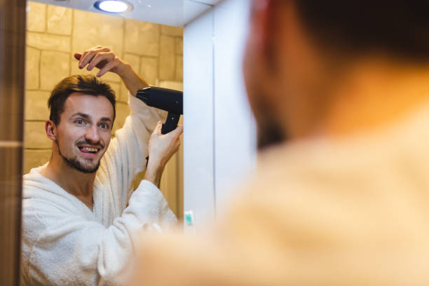 jeune homme souriant en peignoir se séchant les cheveux après avoir pris une douche tout en se regardant dans le miroir de la salle de bain - toothbrush brushing teeth brushing dental hygiene photos et images de collection
