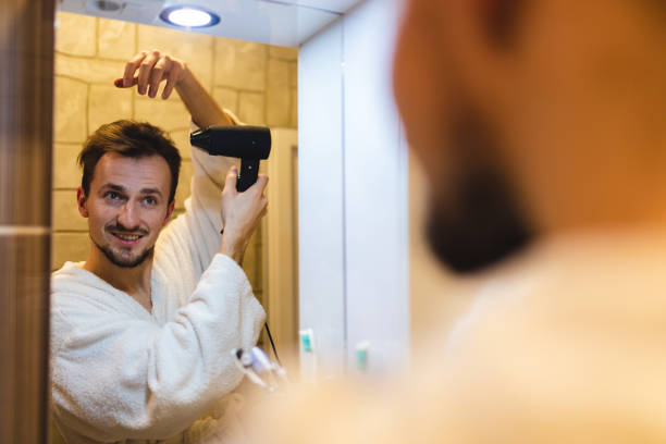 istock Smiling young man in a bathrobe blow drying his hair after taking a shower while looking at himself in the bathroom mirror 1364642556