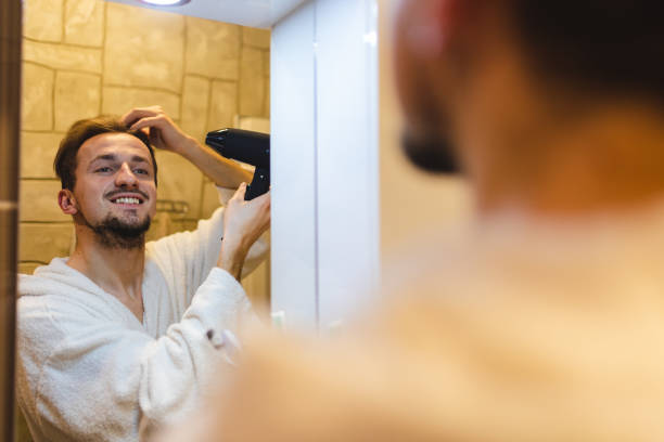 istock Smiling young man in a bathrobe blow drying his hair after taking a shower while looking at himself in the bathroom mirror 1364642598