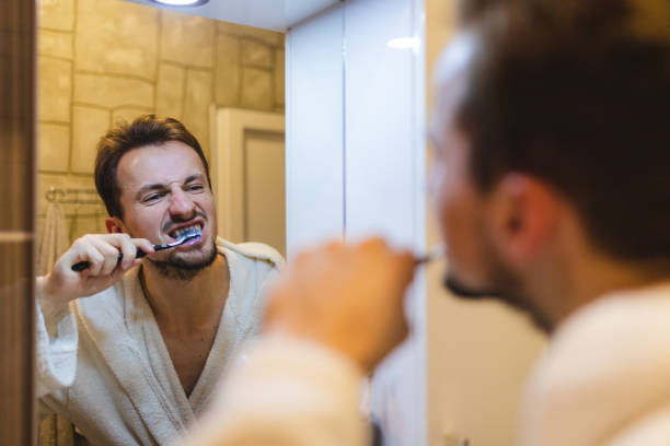 jeune homme en peignoir se brossant les dents tout en se regardant dans le miroir de la salle de bain - toothbrush brushing teeth brushing dental hygiene photos et images de collection
