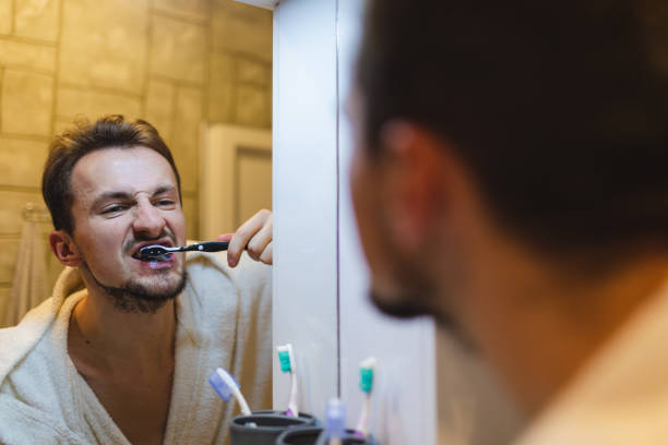 jeune homme en peignoir se brossant les dents tout en se regardant dans le miroir de la salle de bain - brushing teeth photos et images de collection