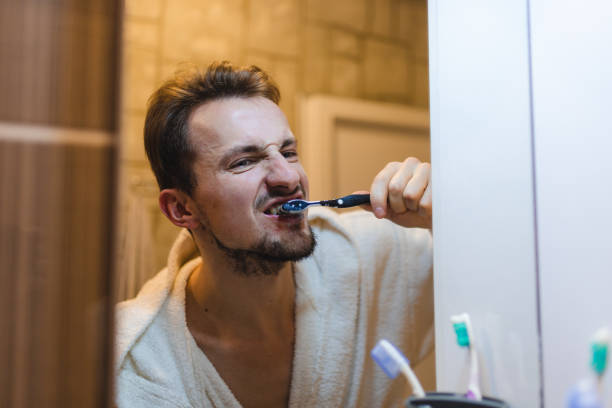 jeune homme en peignoir se brossant les dents tout en se regardant dans le miroir de la salle de bain - toothbrush brushing teeth brushing dental hygiene photos et images de collection