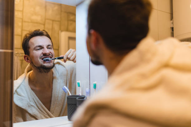 jeune homme en peignoir se brossant les dents tout en se regardant dans le miroir de la salle de bain - brushing teeth photos et images de collection