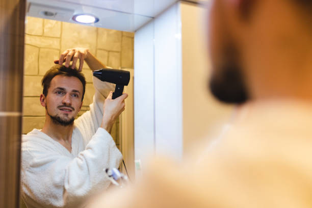 istock Smiling young man in a bathrobe blow drying his hair after taking a shower while looking at himself in the bathroom mirror 1364643224