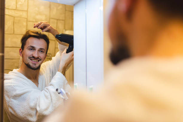 istock Smiling young man in a bathrobe blow drying his hair after taking a shower while looking at himself in the bathroom mirror 1364643226