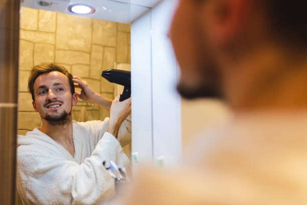 istock Smiling young man in a bathrobe blow drying his hair after taking a shower while looking at himself in the bathroom mirror 1364643234