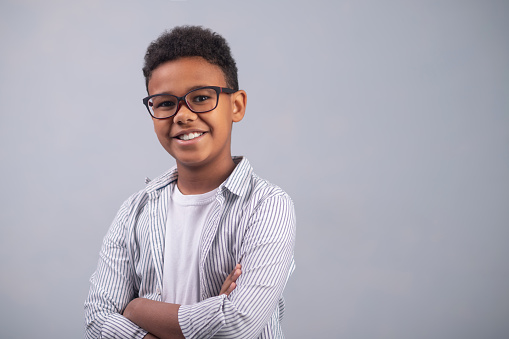 Waist-up portrait of a smiling contented bespectacled kid standing with his arms folded against the white background
