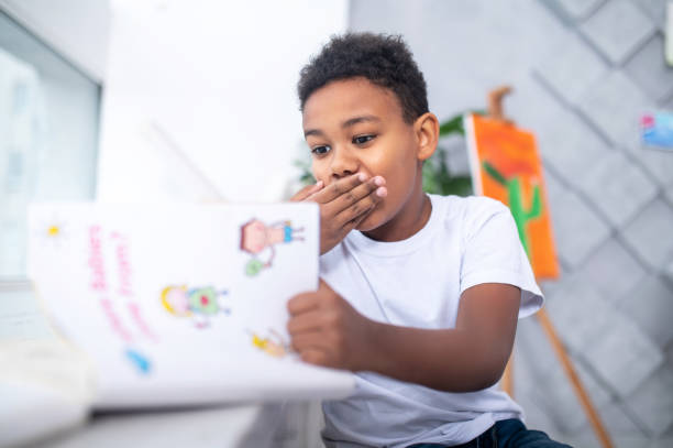 istock Boy looking at book shyly covering mouth with hand 1364643477