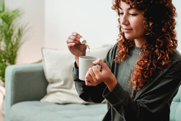 cbd oil dropper - young woman taking cannabis oil in tea drink for anxiety and stress treatment - alternative medicine - druppelfles stockfoto's en -beelden