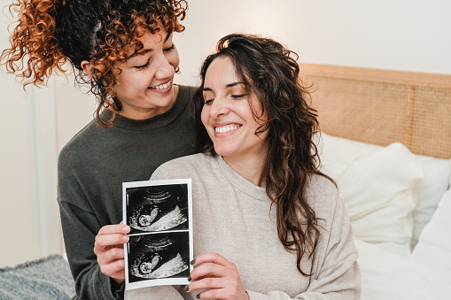 LGBT lesbian couple holding ultrasound photo scan of growing baby in pregnancy time - Focus on right woman face