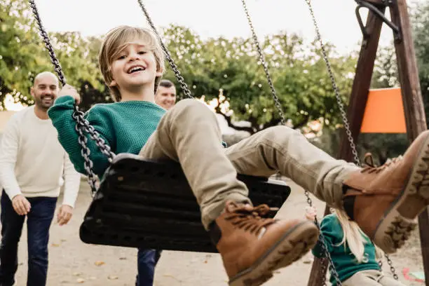 Photo of LGBT family - Happy children sons and fathers having fun swinging on swing at city park - Focus on left kid face