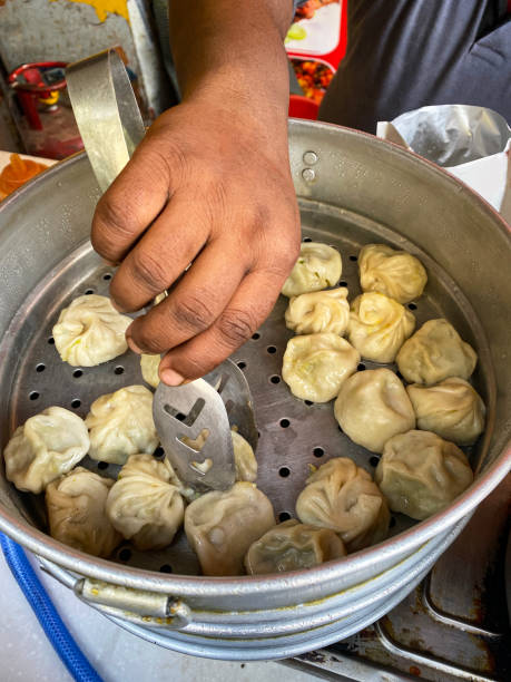 Stock photo showing a close-up view of a street food meal of steaming dumplings (Momos) in a stainless steel steamer being server by an unrecognisable street vendor with metal tongs into a takeaway packet. imagen de un vendedor amrococible de comida callejera india que usa pinzas para servir para mover momos (albóndigas del sur de asia), harina blanca y masa de agua llena de verduras mezcladas, desde un vaporizador de acero inoxidable hasta un paquete para