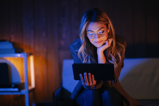 Beautiful smiling woman sitting on the couch, leaning forward, holding a tablet and reading something. She is wearing eyeglasses. It is dark in the room and her face is illuminated from the screen.