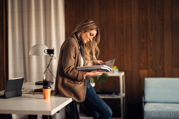 Beautiful Businesswoman Sitting on her Desk Holding Big Books Pretty woman sitting on her desk. She is holding two big catalogues or books and she is looking down at the photos. There is a disposable orange paper cup for coffee and tablet on the desk. catalogue stock pictures, royalty-free photos & images