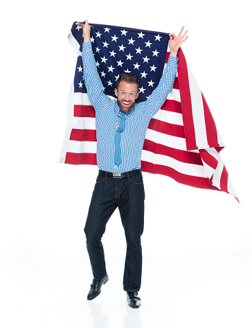 Full length of aged 30-39 years old caucasian male standing in front of white background wearing button down shirt who is shouting and celebrating and showing patriotism and holding american flag