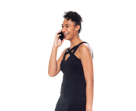 Side view of aged 20-29 years old who is beautiful african ethnicity young women standing in front of white background wearing dress who is talking and using mobile phone