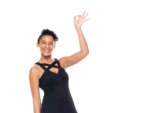 Front view of aged 20-29 years old who is beautiful african ethnicity young women standing in front of white background wearing dress who is smiling and showing hand raised